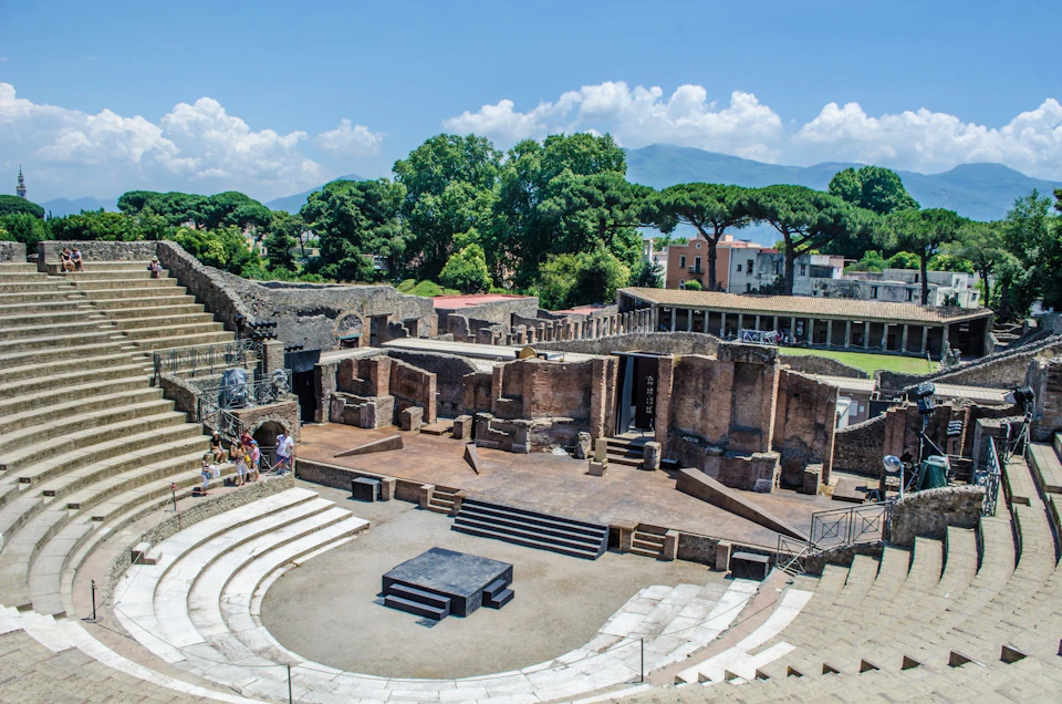 Ancient amphitheater with stone seating, central stage, and surrounding ruins. Trees and mountains in the background.