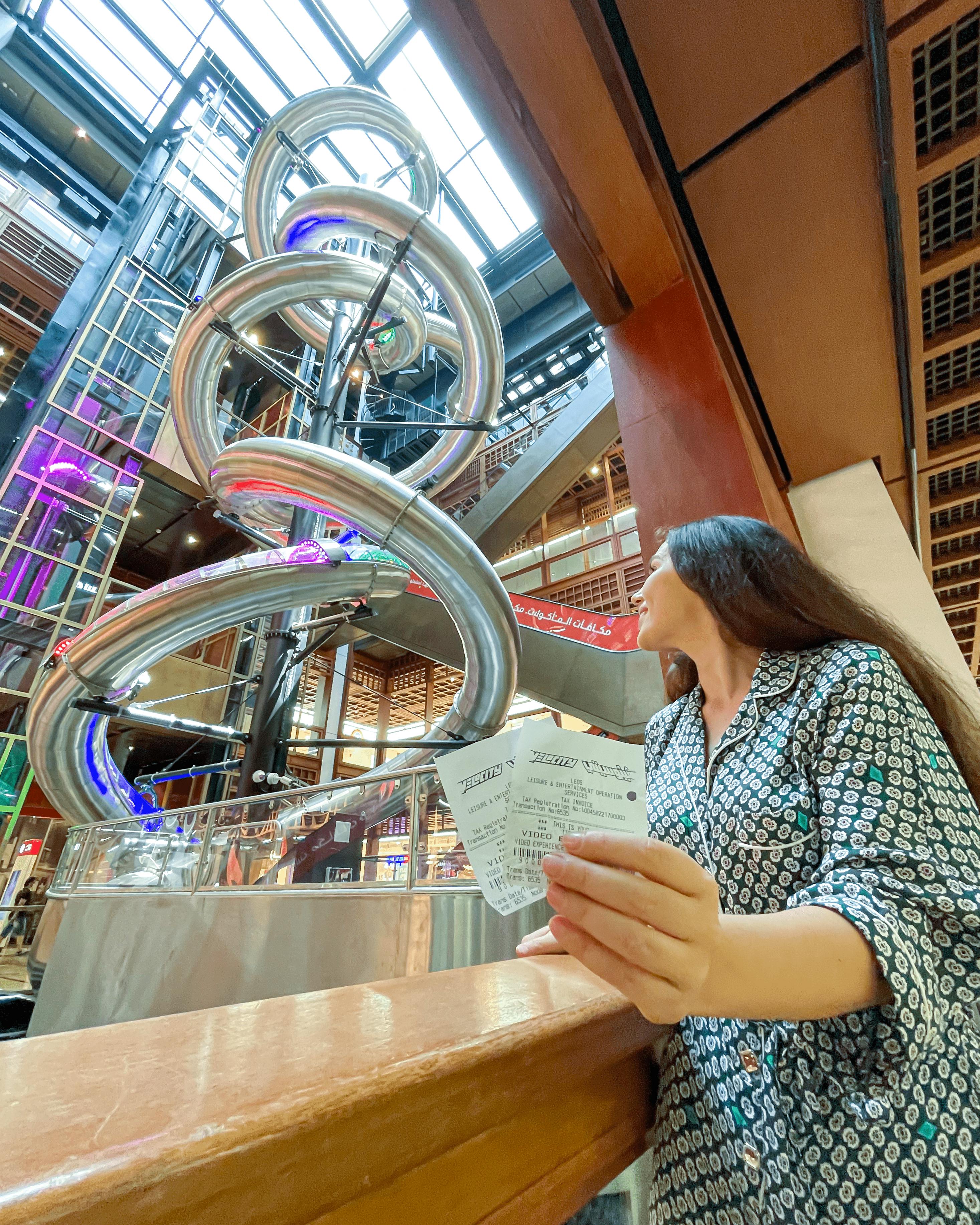 Žena drží dva lístky a dívá se na velkou spirálovou skluzavku uvnitř moderní vícepodlažní budovy.