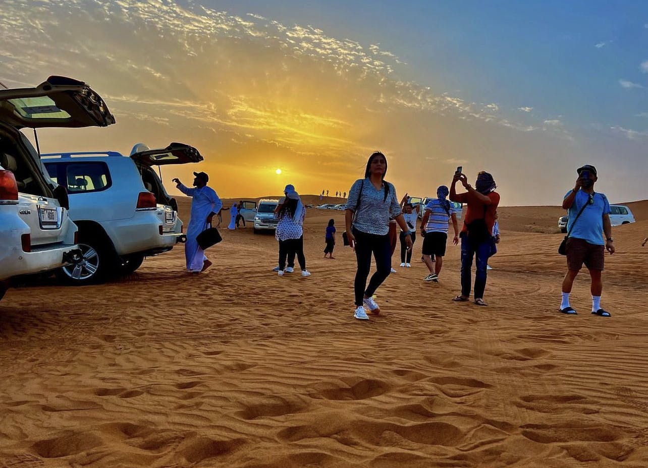 People walking on desert sand near parked SUVs at sunset, with others taking photos and the bright sun low on the horizon.