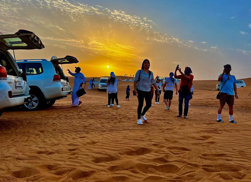People walking on desert sand near parked SUVs at sunset, with others taking photos and the bright sun low on the horizon.