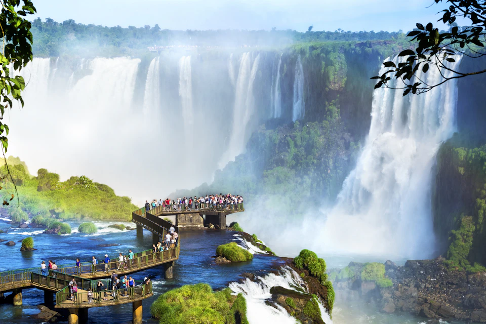 Tourists stand on elevated walkways with panoramic views of large, mist-filled waterfalls cascading down lush green cliffs.