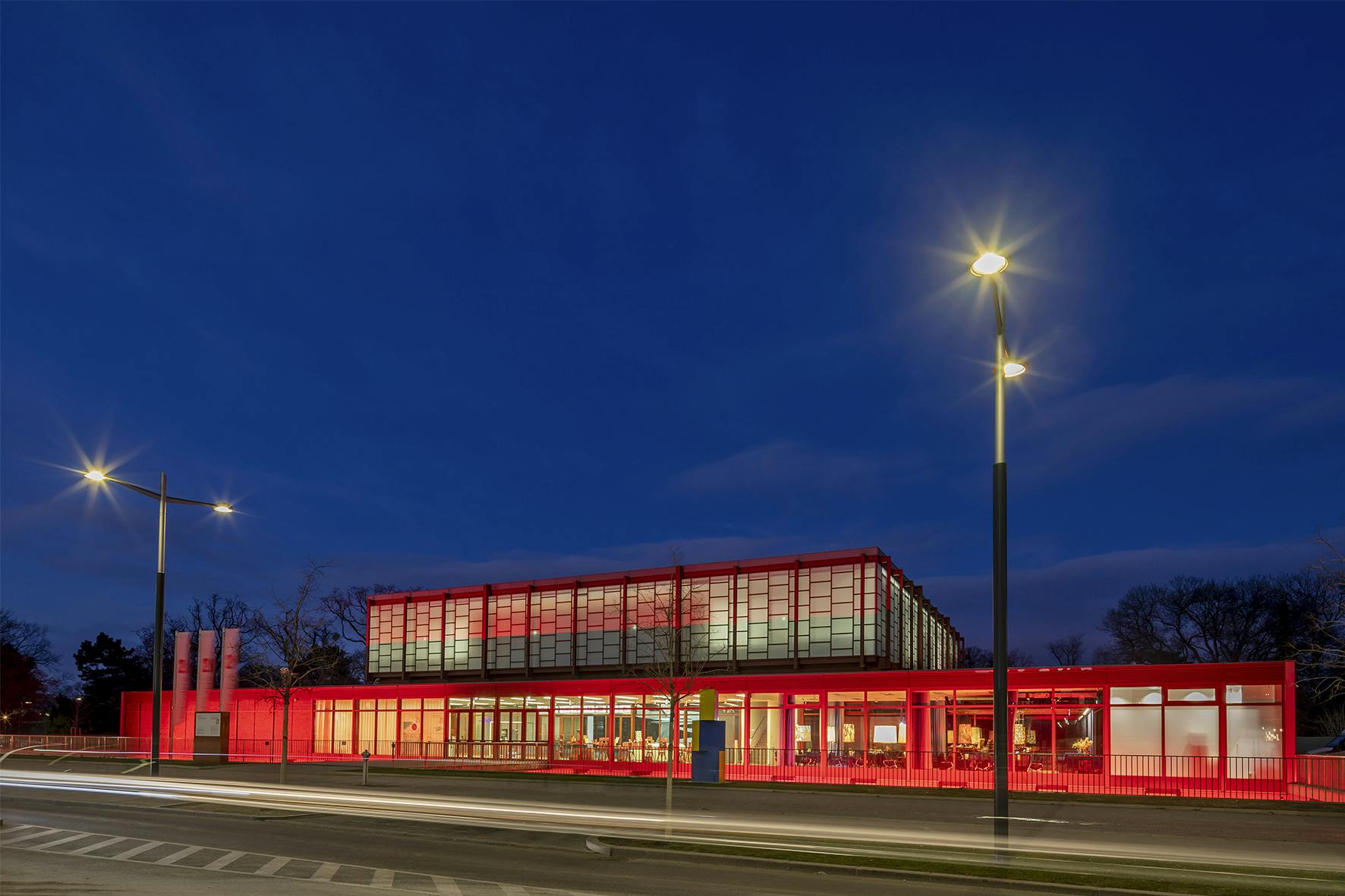 Ένα μοντέρνο κτίριο με γυάλινη επένδυση που φωτίζεται με κόκκινο χρώμα τη νύχτα με φώτα του δρόμου και φώτα αυτοκινήτων με θολή κίνηση στο προσκήνιο.