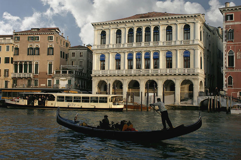 A gondolier rows a gondola on a canal in front of a large ornate building, with a water bus and other buildings in the background.