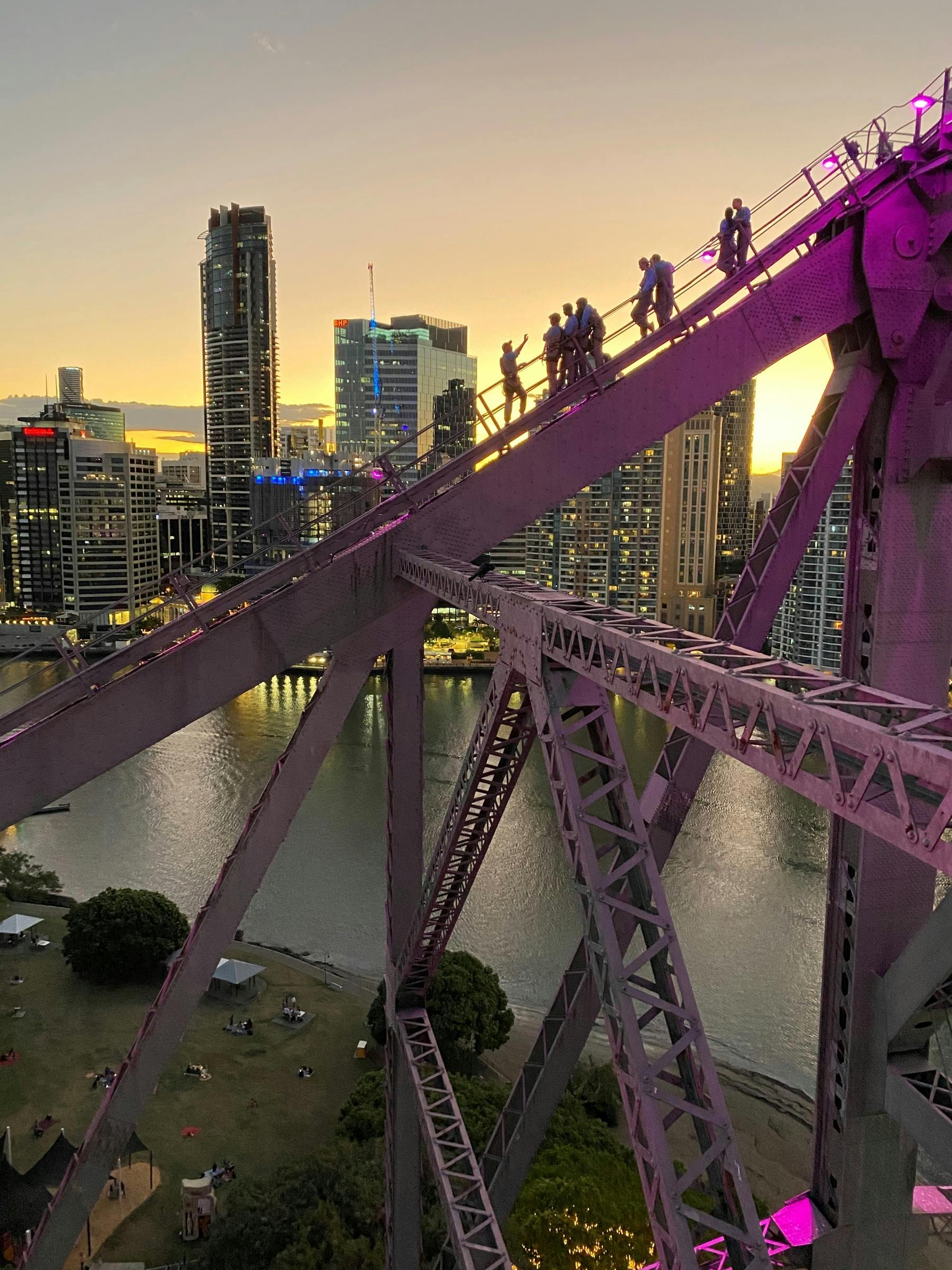 해가 질 무렵 도시의 스카이라인과 강을 배경으로 보라색 다리를 걷는 사람들.