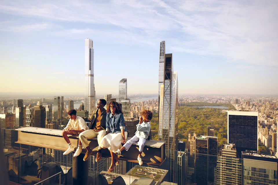 A group of four people sitting on a high beam overlooking a cityscape with tall buildings and a park.
