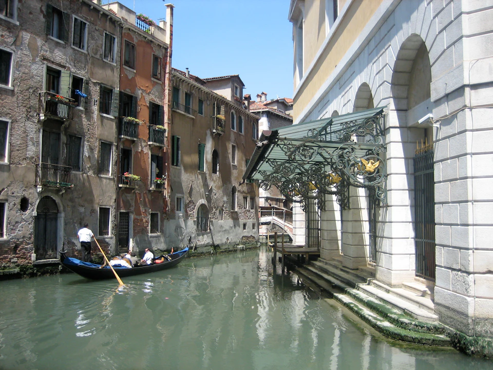 A gondola navigates a narrow canal between aged buildings and a grand entrance in Venice, Italy, under a clear sky.