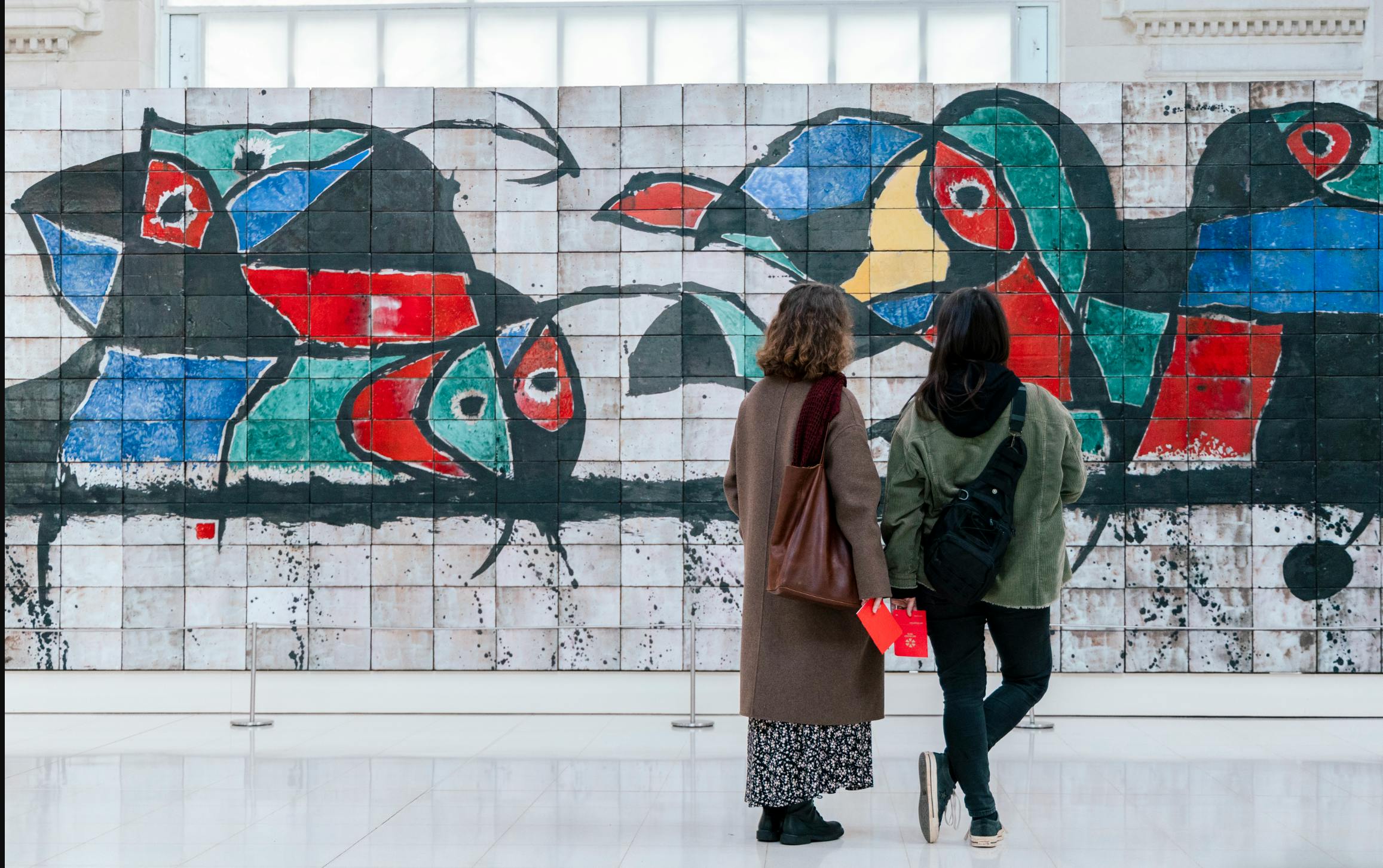 Dos individus es paren davant d'un gran mural abstracte compost per formes i patrons de colors sobre una superfície de rajoles.