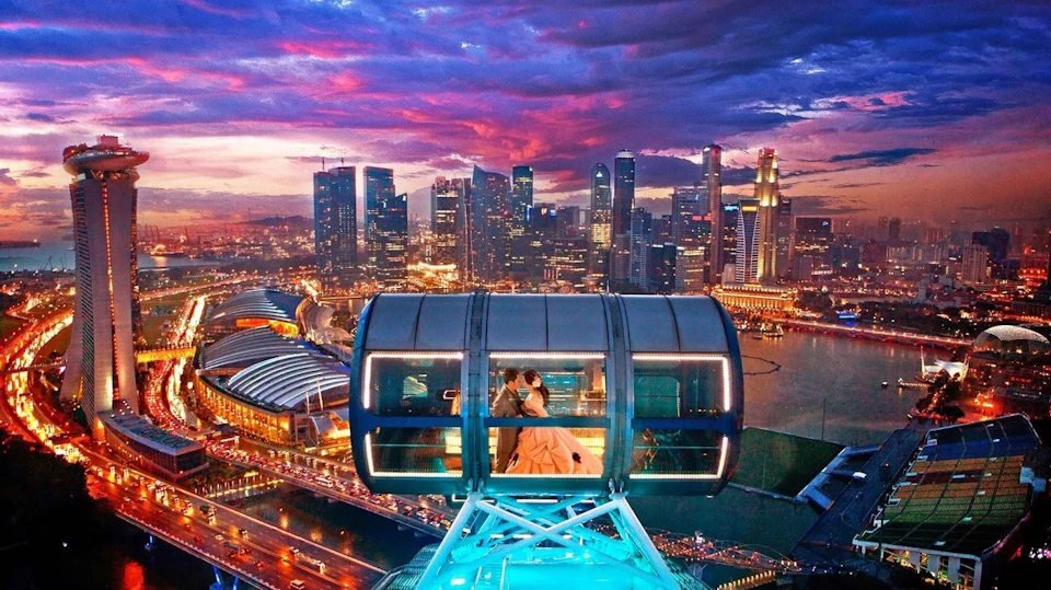 A couple in formal attire sits inside a glass observation pod during sunset, overlooking a cityscape with illuminated skyscrapers.
