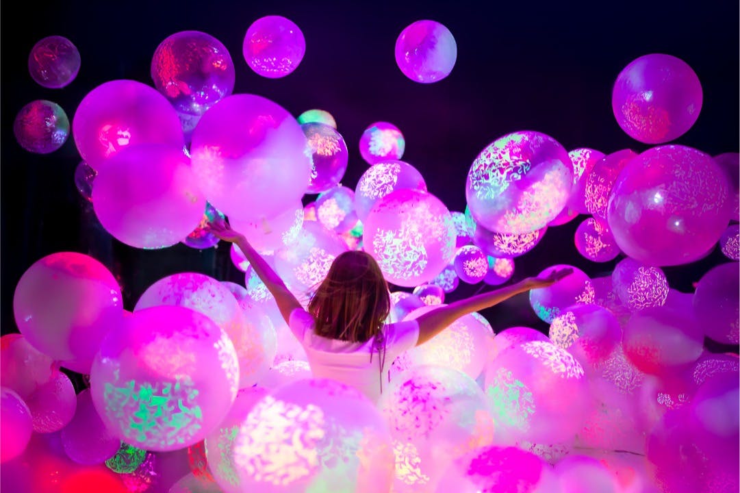 Une personne entourée de ballons lumineux et colorés sur un fond sombre, les bras levés dans une posture joyeuse.