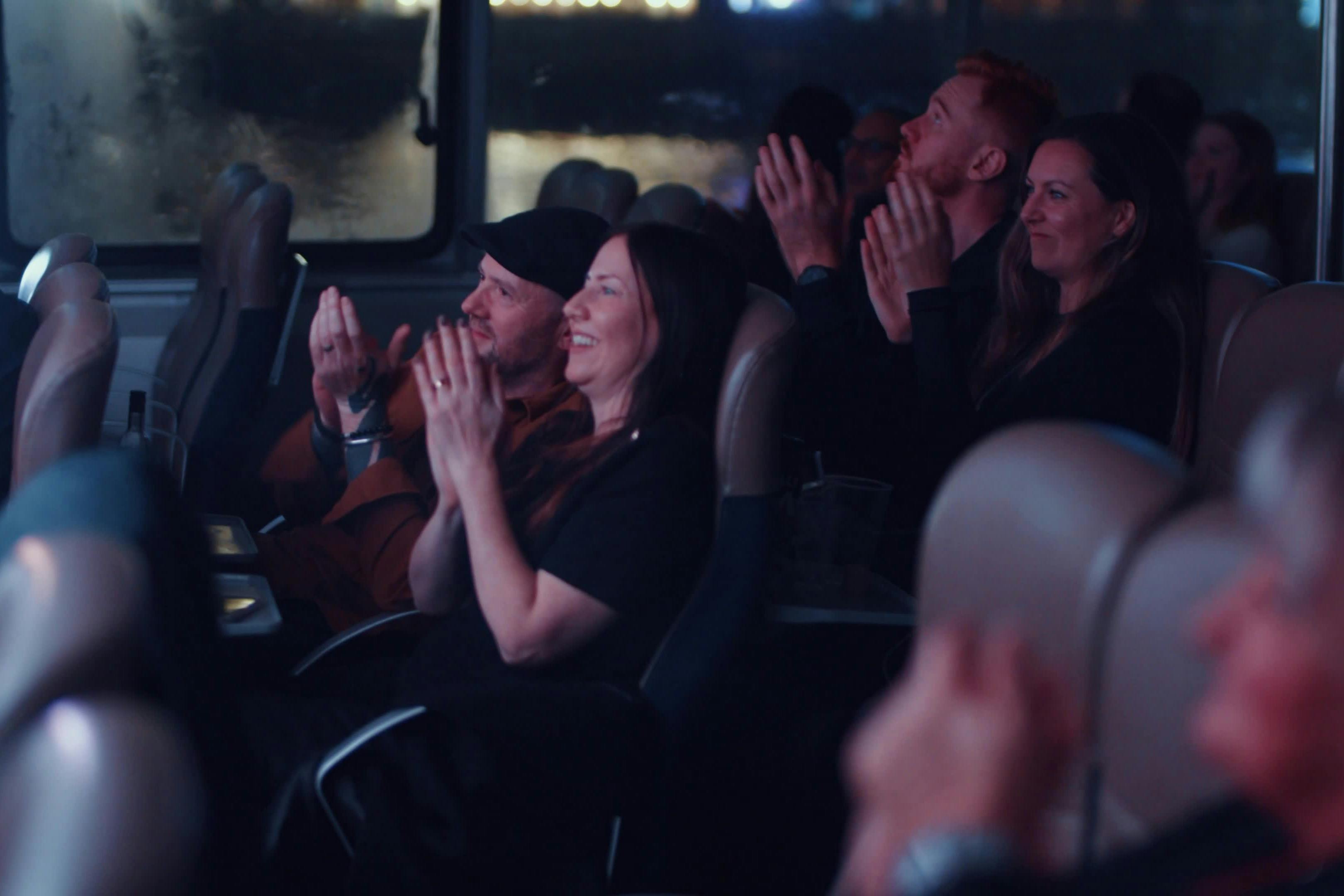 A group of people sitting in a theater clapping and smiling during a performance, with dim lighting and blurred background.