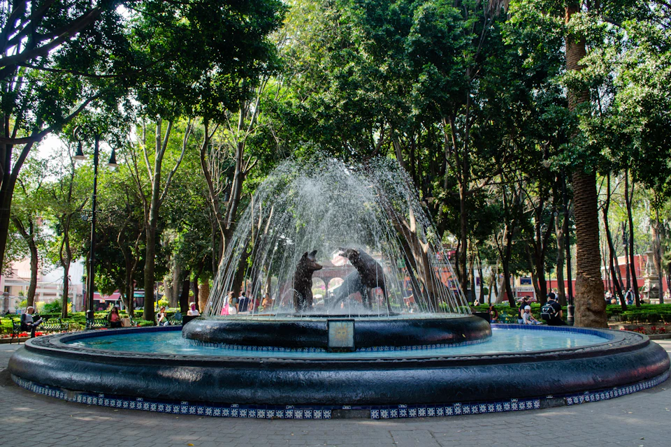 A fountain in a park with statues of two animals fighting in the center, surrounded by trees and people sitting nearby.