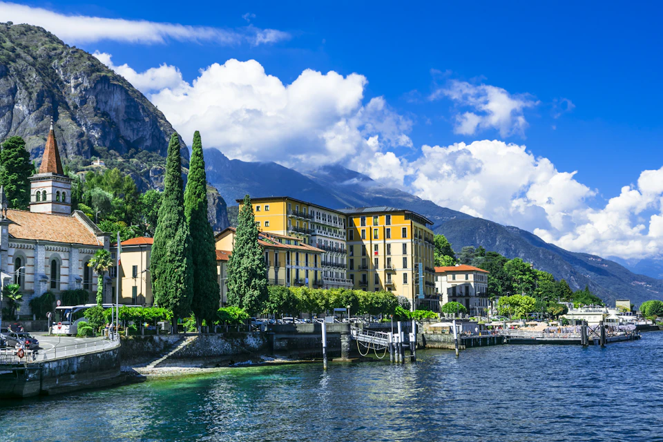 Historic yellow building on lakeshore, surrounded by trees, with mountains and blue sky with clouds in the background.