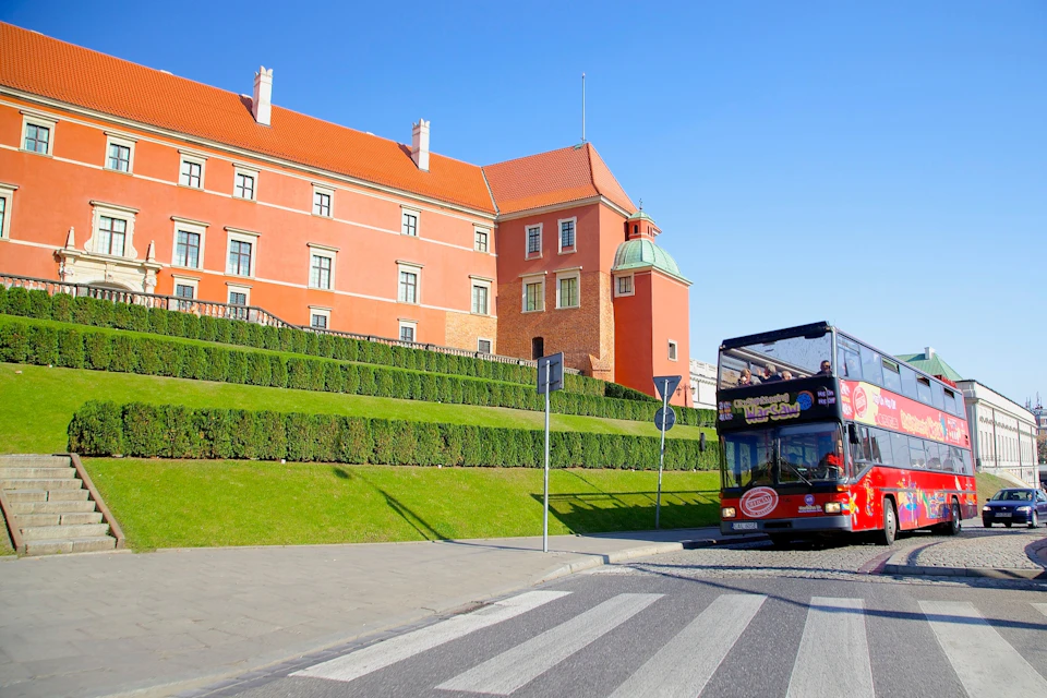 A double-decker tour bus passes by a historical red building with green hedges in front on a sunny day.