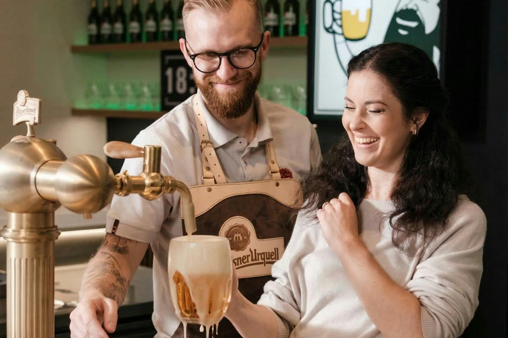 Ένας άντρας και μια γυναίκα χαμογελούν καθώς ο άντρας χύνει μπύρα από μια βρύση σε ένα ποτήρι. Η γυναίκα κρατάει το ποτήρι κάτω από τη βρύση.