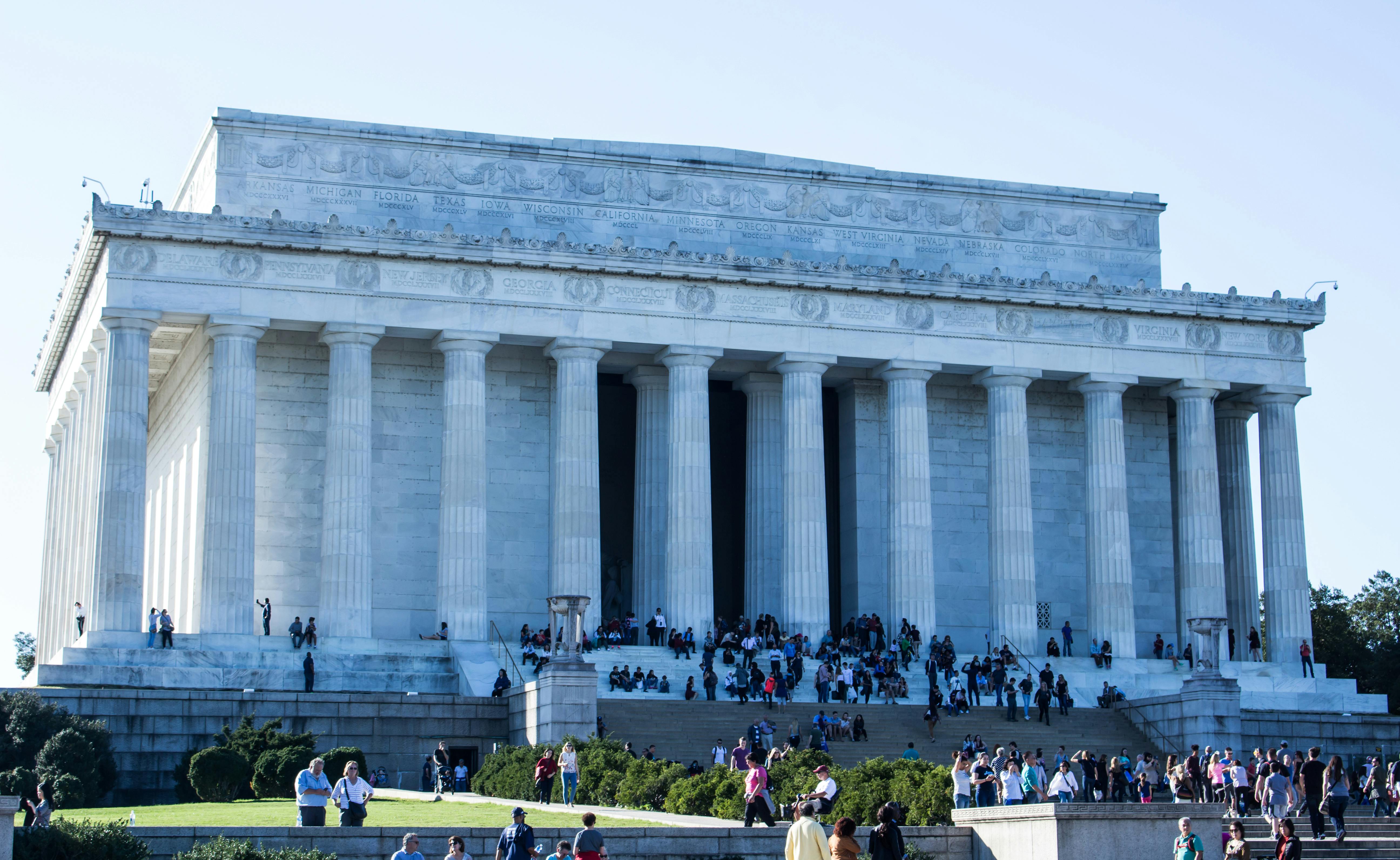 Numerous people gather on steps and at the base of the Lincoln Memorial, a large marble building with tall columns.