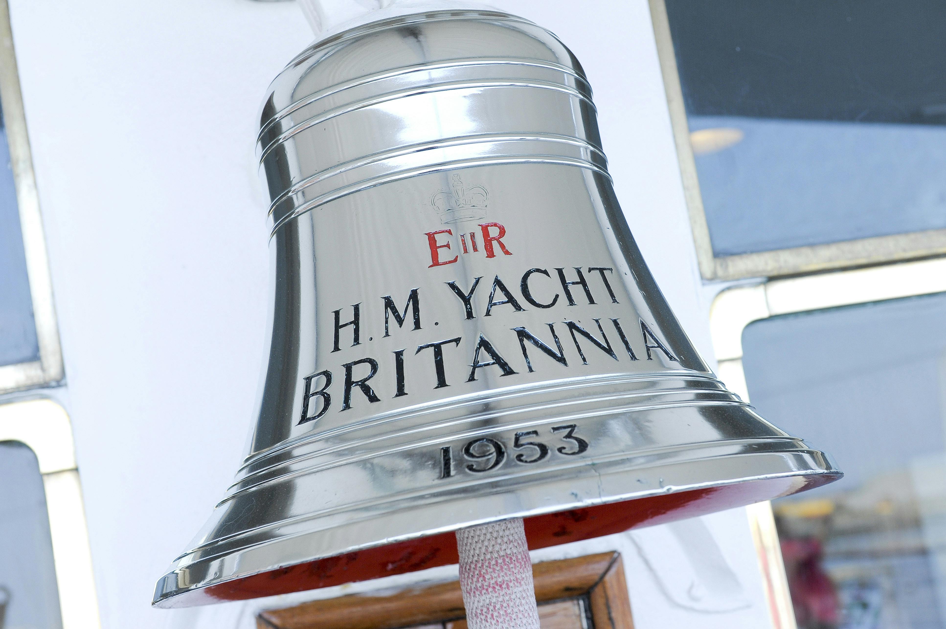 Una campana lucida con la scritta "H.M. YACHT BRITANNIA", "1953" e i segni della corona è appesa vicino a una finestra.