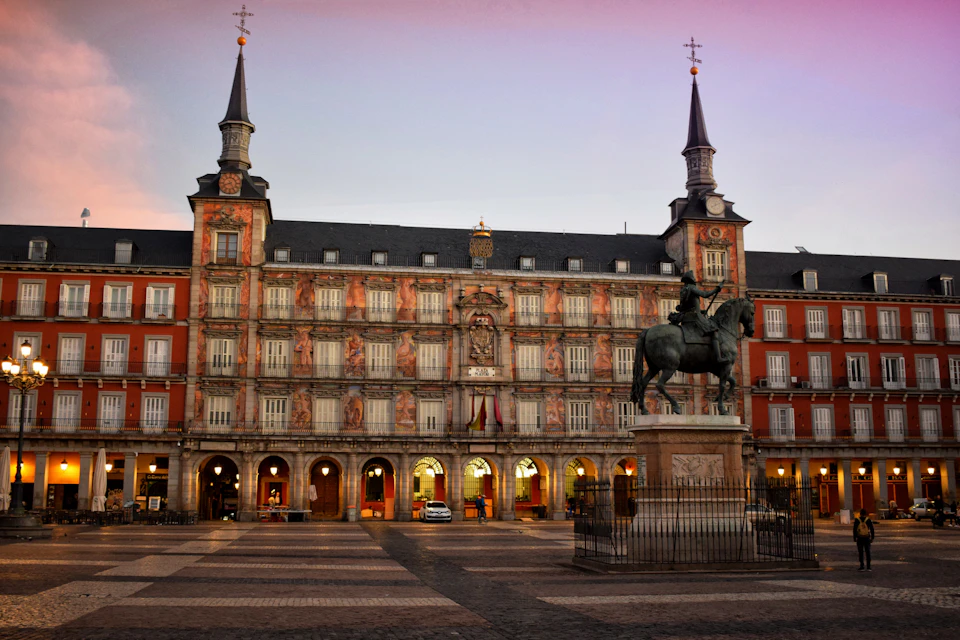 A historic square at sunset with an ornate building, featuring two spires and a statue of a rider on horseback in the foreground.