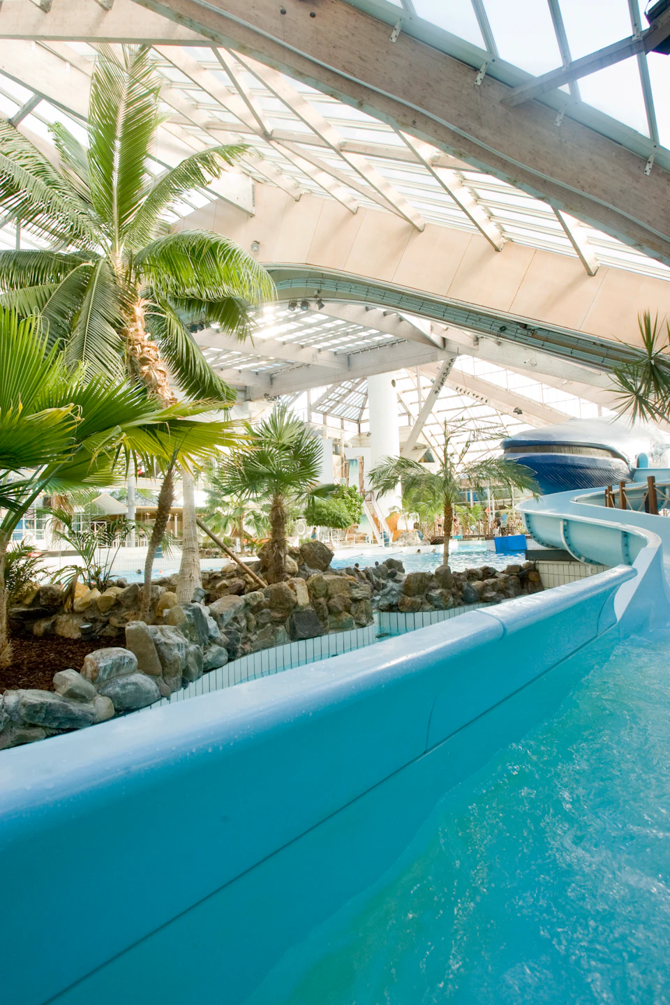 Indoor water park with a tropical theme, featuring a blue water slide, palm trees, rocks, and a large glass roof.