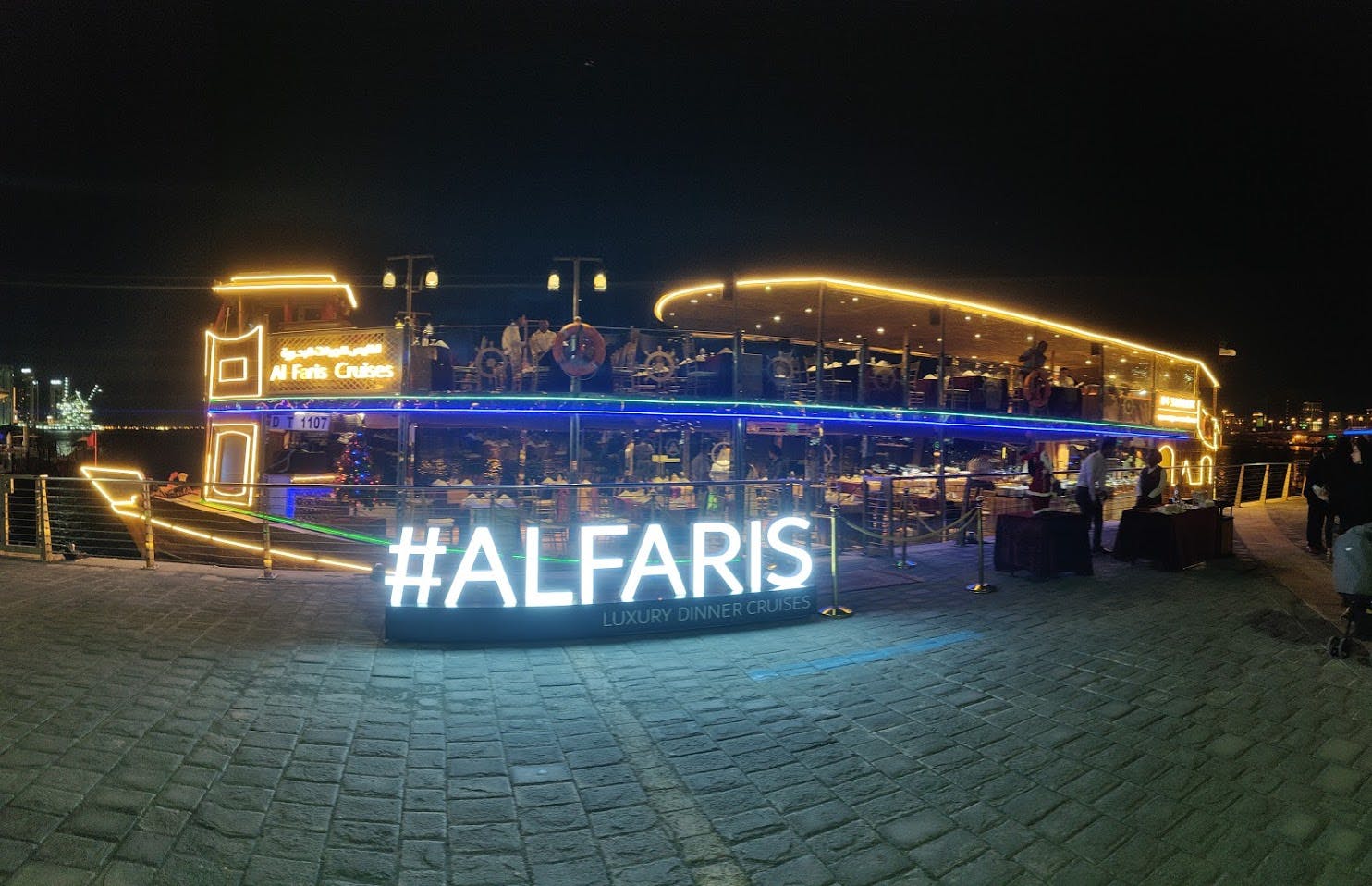 A brightly lit boat with "ALFARIS Luxury Dinner Cruises" sign, docked at night with decorations and people visible on board.