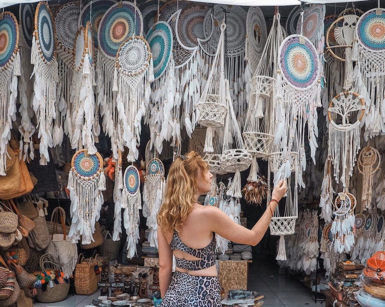 A person in a patterned outfit looks at a display of colorful dreamcatchers and macramé items in a shop.