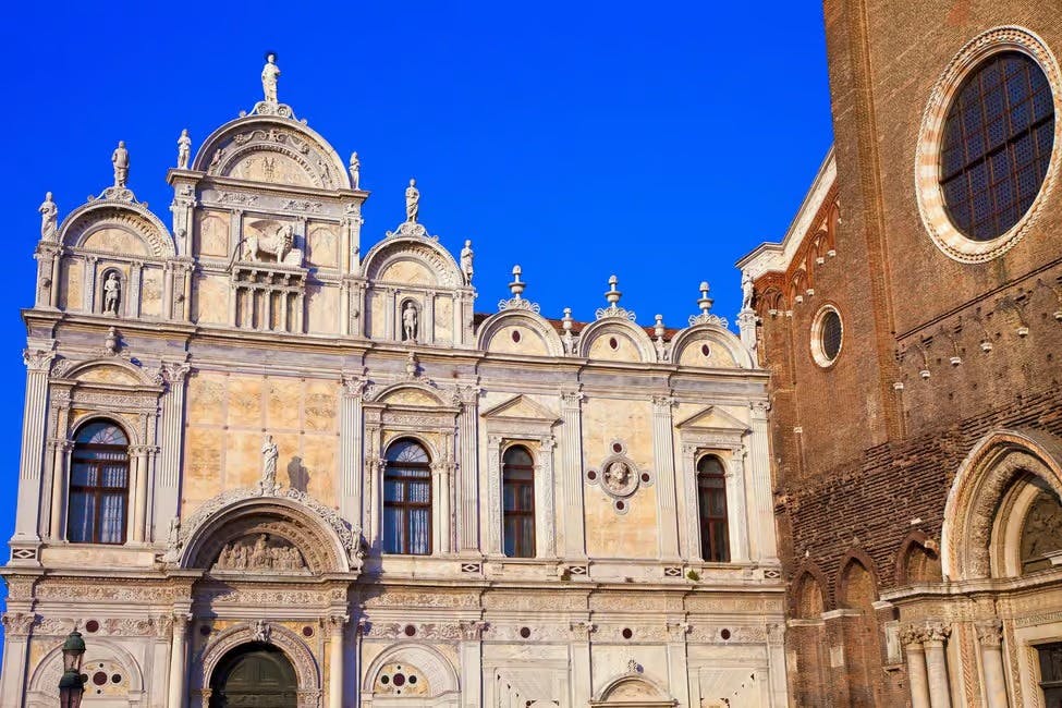 Um edifício histórico de pedra branca com detalhes arquitectónicos ornamentados sob um céu azul claro, adjacente a uma estrutura de tijolo.