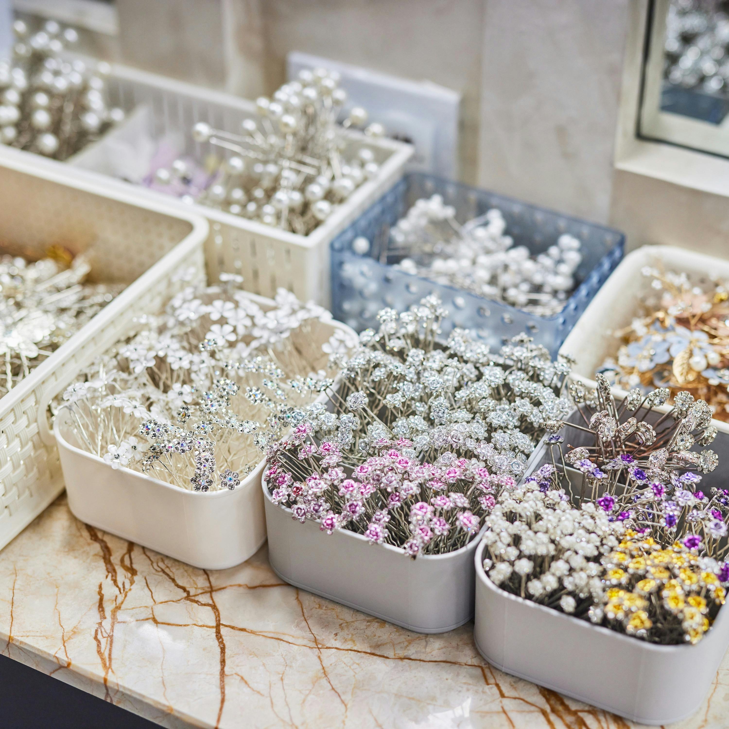 Assorted decorative floral pins in various containers on a marble surface, featuring white, pink, and metallic hues.