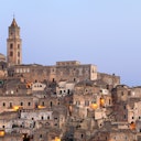 An ancient hillside town with tightly packed stone buildings and a tall bell tower under a clear evening sky.