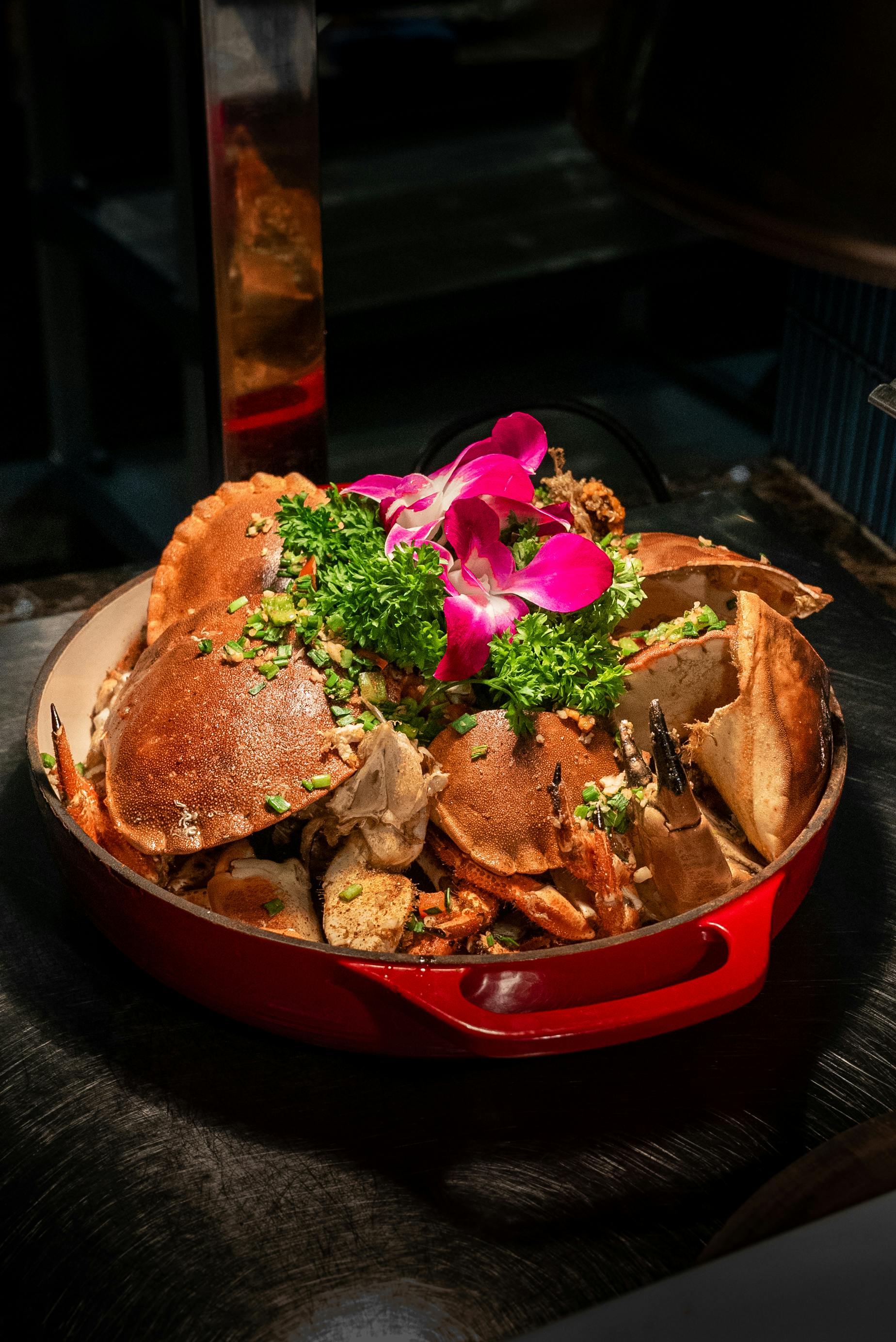 A red dish containing cooked whole crabs garnished with parsley, chopped vegetables, and a purple orchid on top.