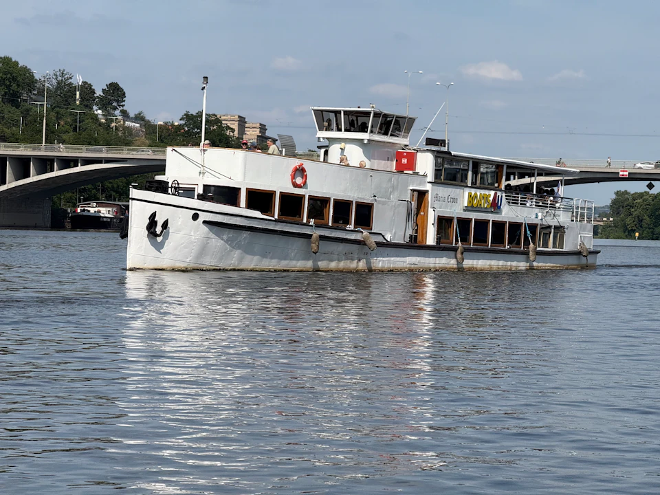 A white passenger boat on a calm river under a clear sky, with trees and buildings in the background.