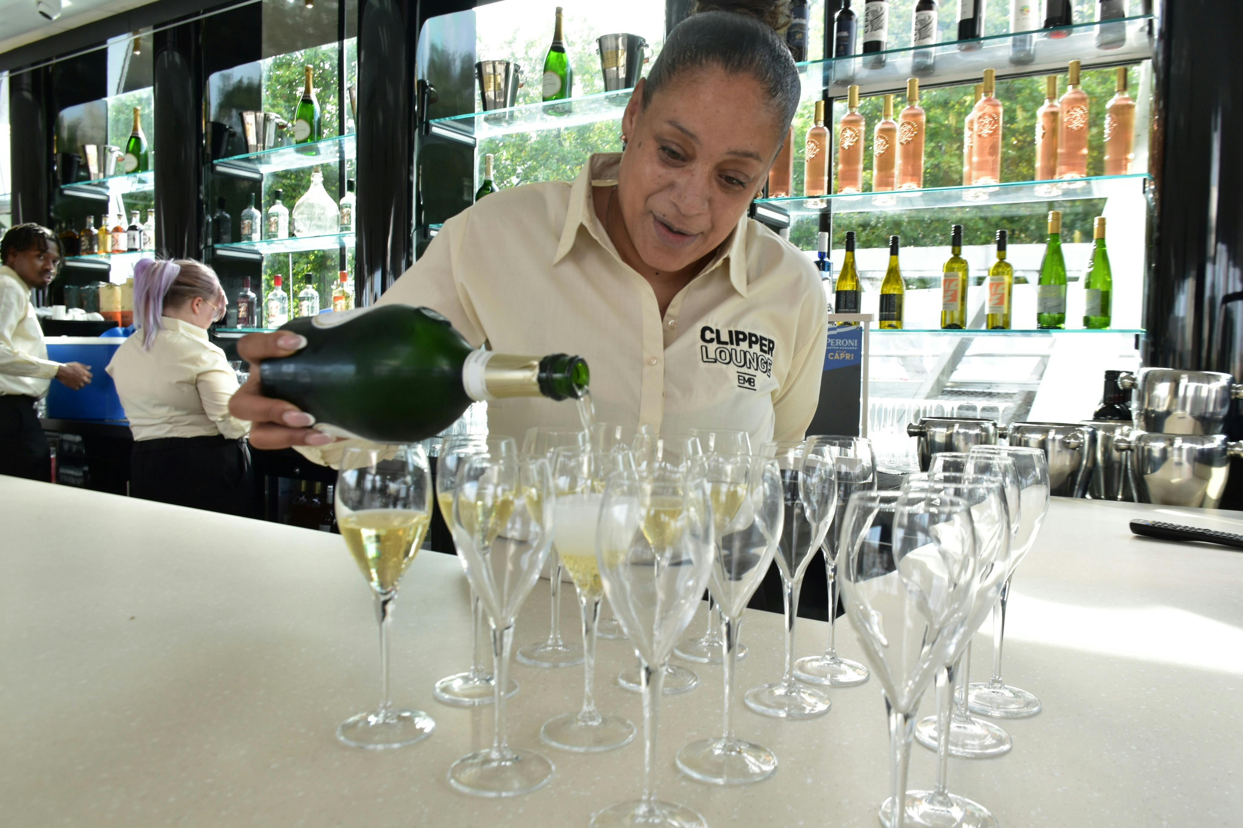 バーで複数のグラスにシャンパンを注ぐ女性。背景にはアルコールボトルが表示されている。