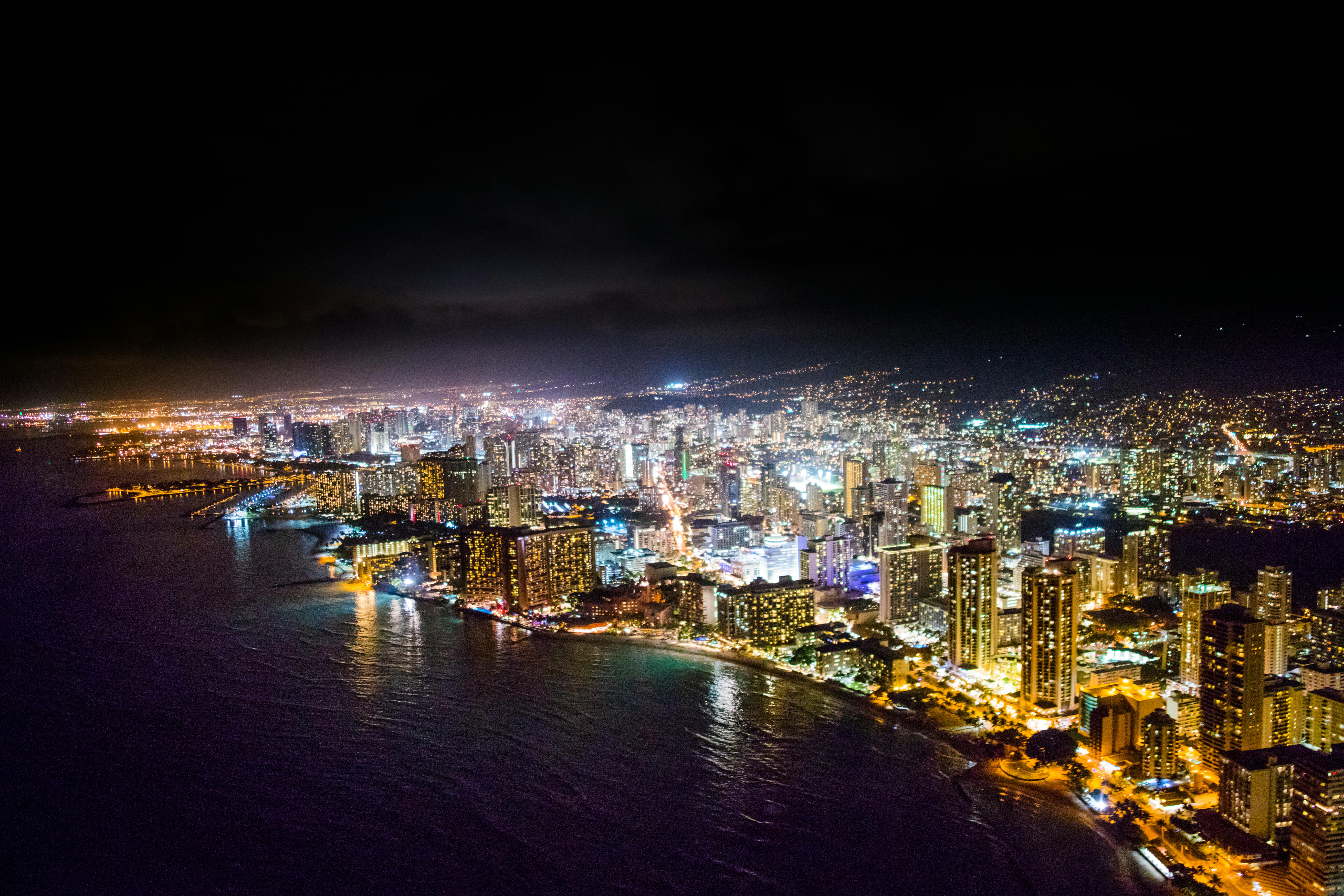Aerial view of a coastal city at night, brightly lit buildings and streets, with dark water and a black sky.