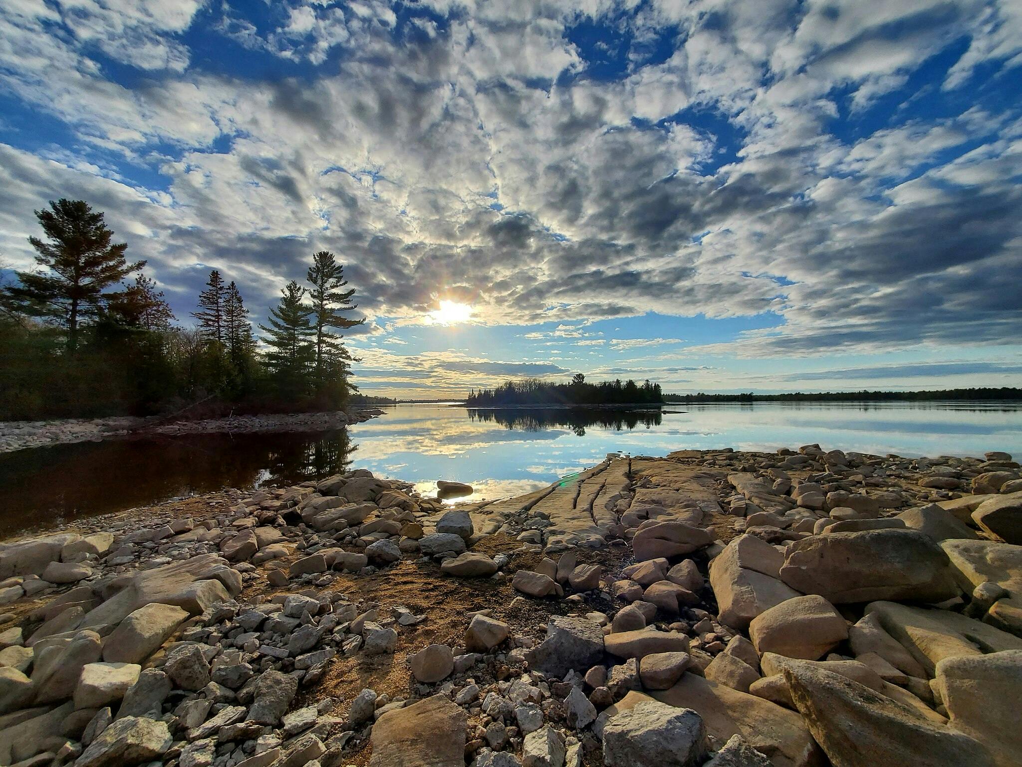 Rocky shoreline with scattered boulders leading to a calm lake, reflecting a partly cloudy sky and setting sun, framed by trees.