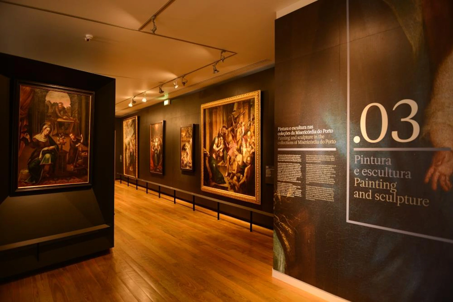 Art gallery with framed paintings on dark walls, informational text on the right, and a wooden floor under track lighting.