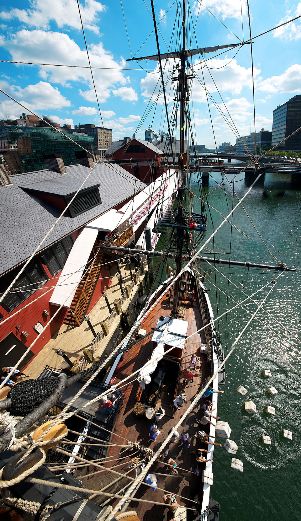 Bird's-eye view of a docked ship with intricate rigging next to a red wooden building in a city with bridges in the background.