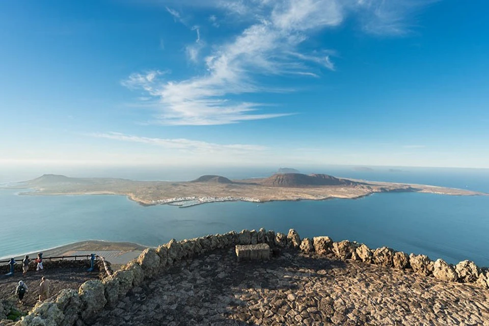 Rocky lookout point with a stone bench overlooking a coastline, distant mountains, and a clear, blue sky with wispy clouds.