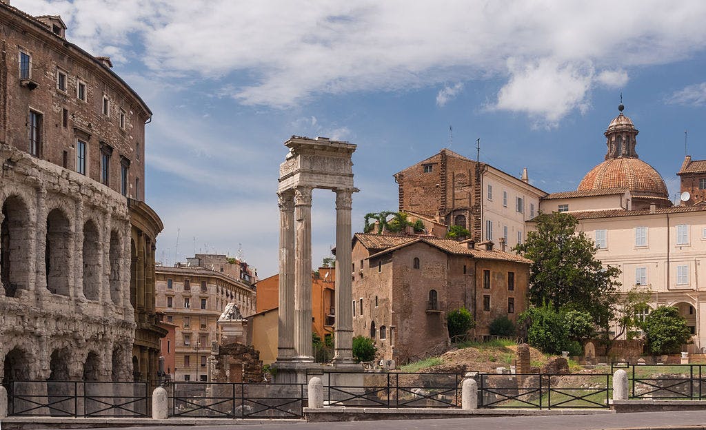 Antiche rovine romane, tra cui una grande struttura ad arco e una singola colonna eretta, con edifici storici e una cupola sullo sfondo.