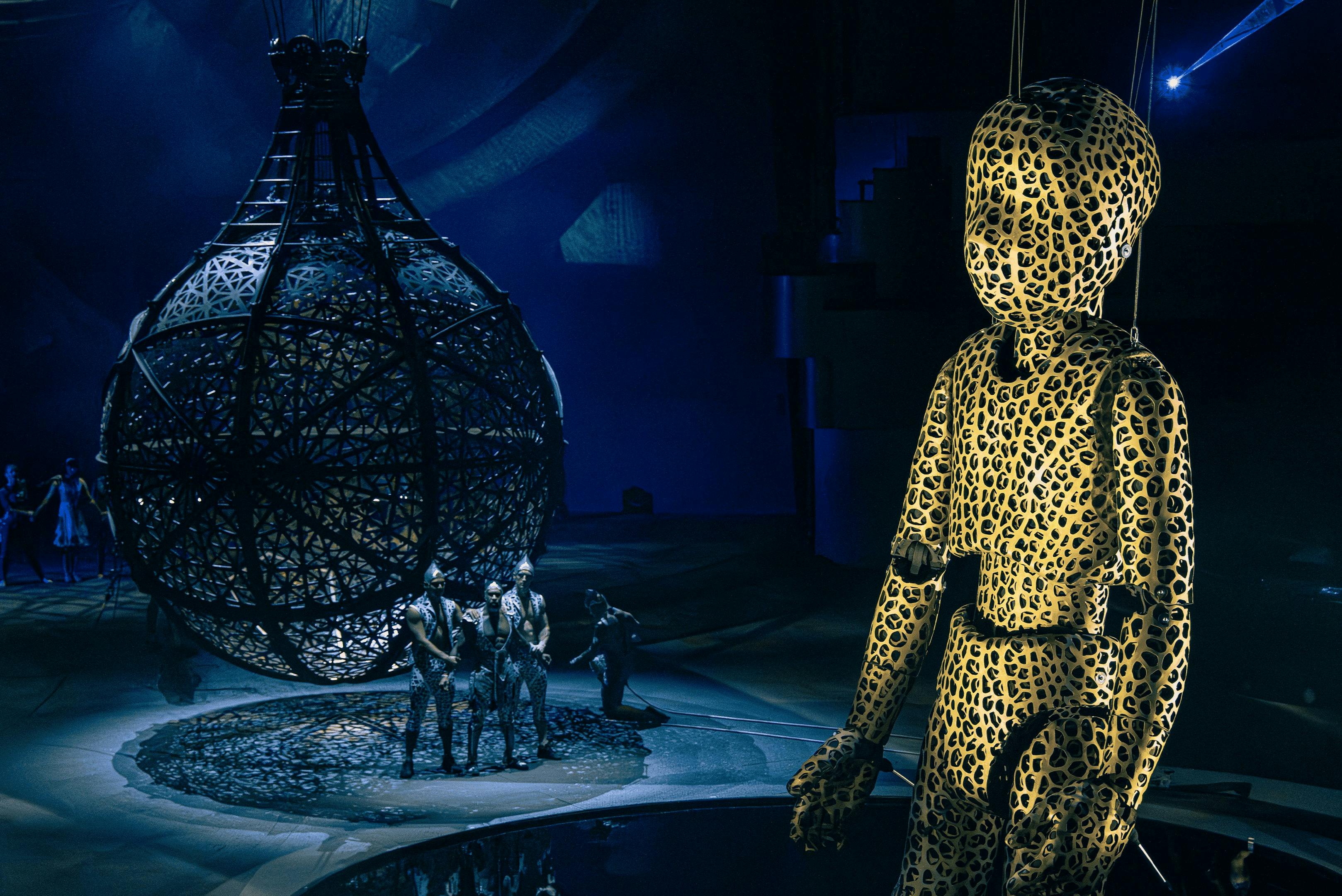 Een dramatische scène met een grote mannequin met luipaardmotief, een bolvormige kooi en artiesten in ingewikkelde kostuums.