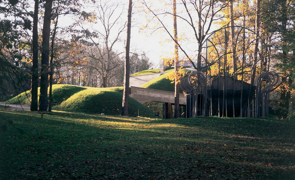 A modern building with grass-covered, hill-like roofs, surrounded by trees with autumn leaves in a wooded area.