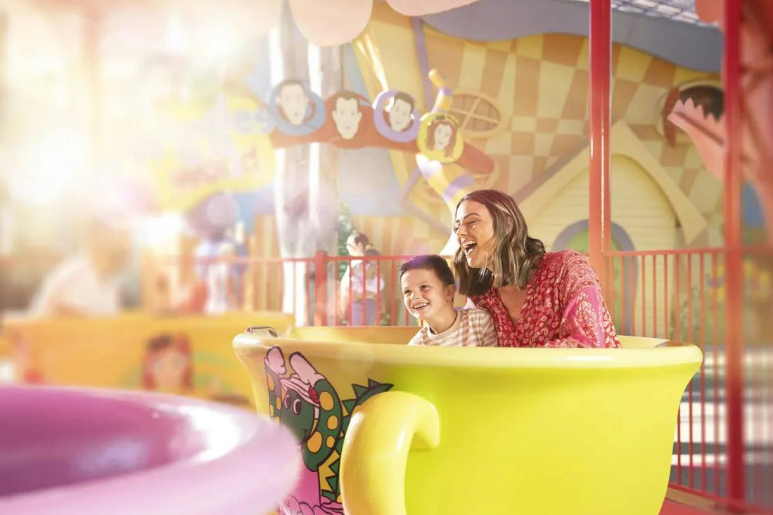 A smiling woman and a child riding a yellow teacup amusement ride in a colorful, whimsical setting.