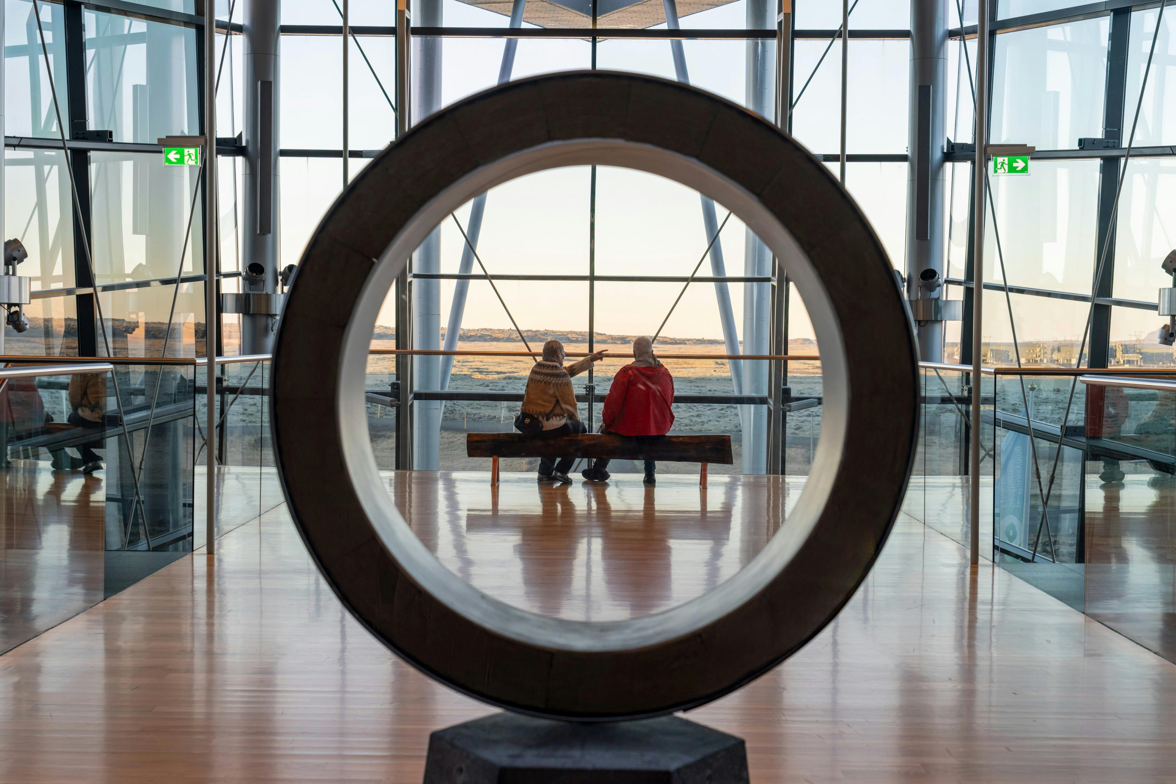 Zwei Personen sitzen auf einer Bank in einem modernen Gebäude, eingerahmt von einer großen runden Skulptur, mit Blick auf eine ferne Landschaft.