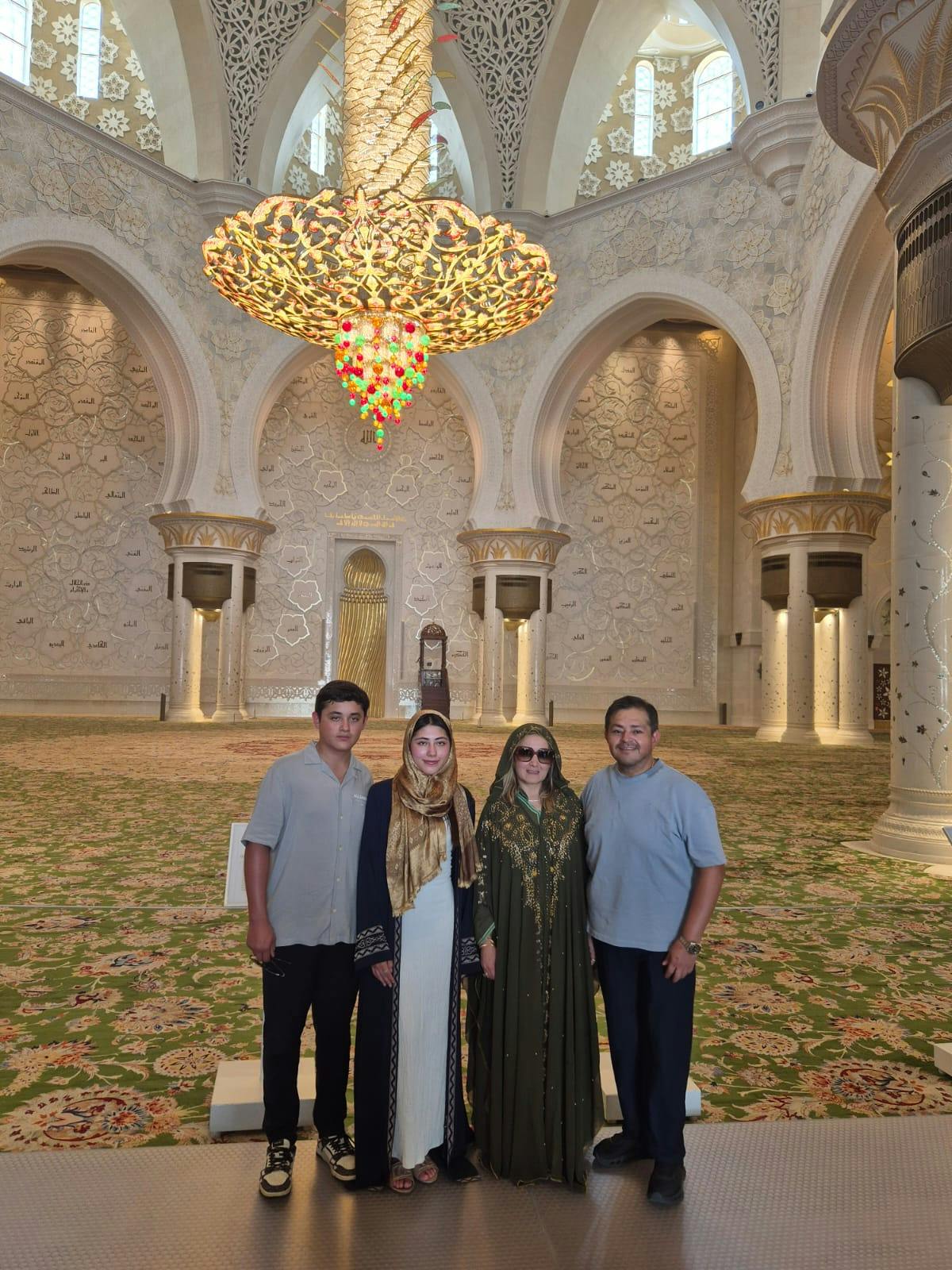 Four people stand inside an ornate mosque with detailed arches, large columns, and an elaborate chandelier above them.