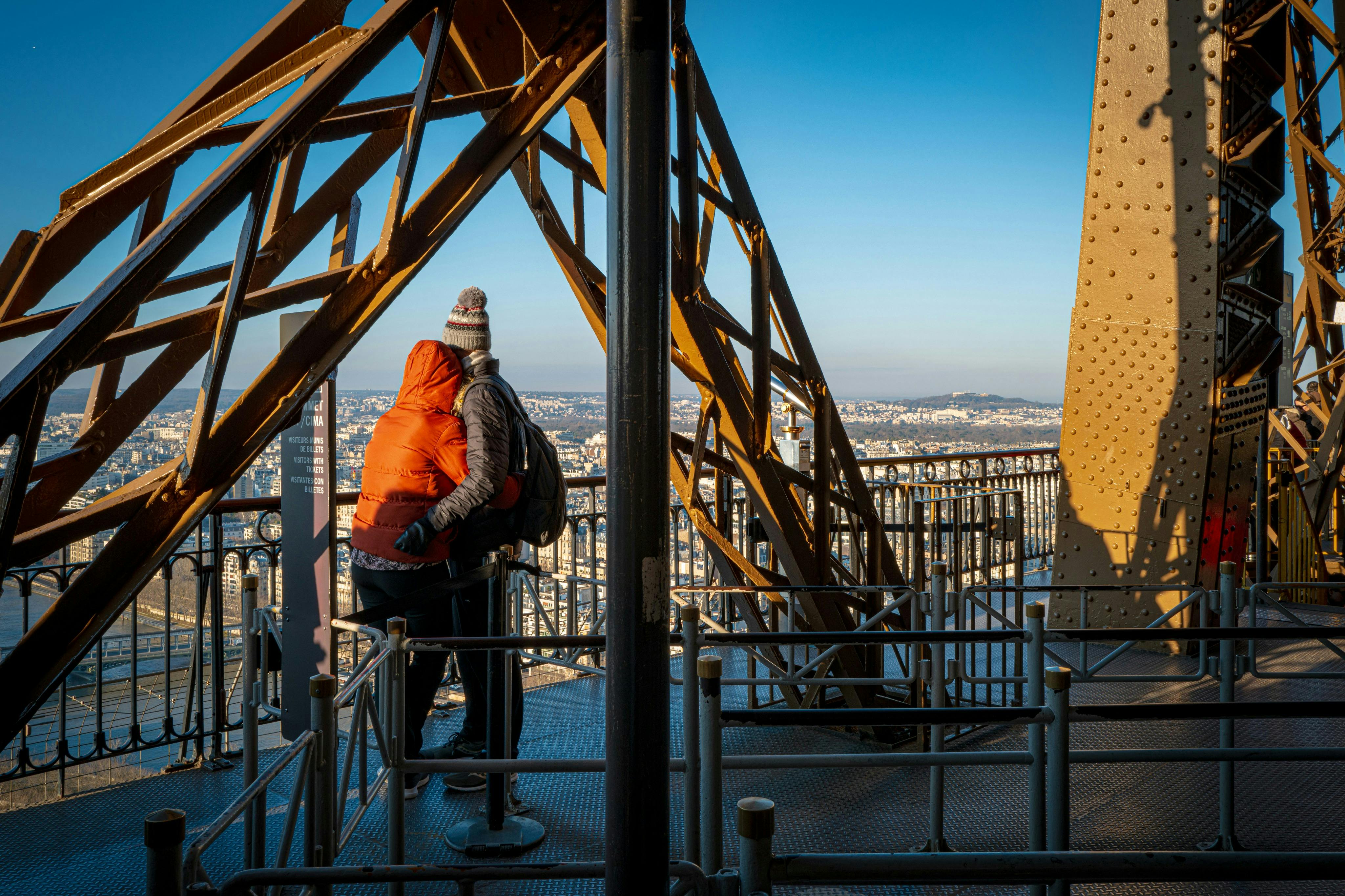 两人站在埃菲尔铁塔的平台上，四周环绕着金属横梁和栏杆，俯瞰城市景观。