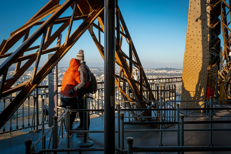 Two people stand on a platform of the Eiffel Tower, surrounded by metal beams and railings, overlooking a cityscape.