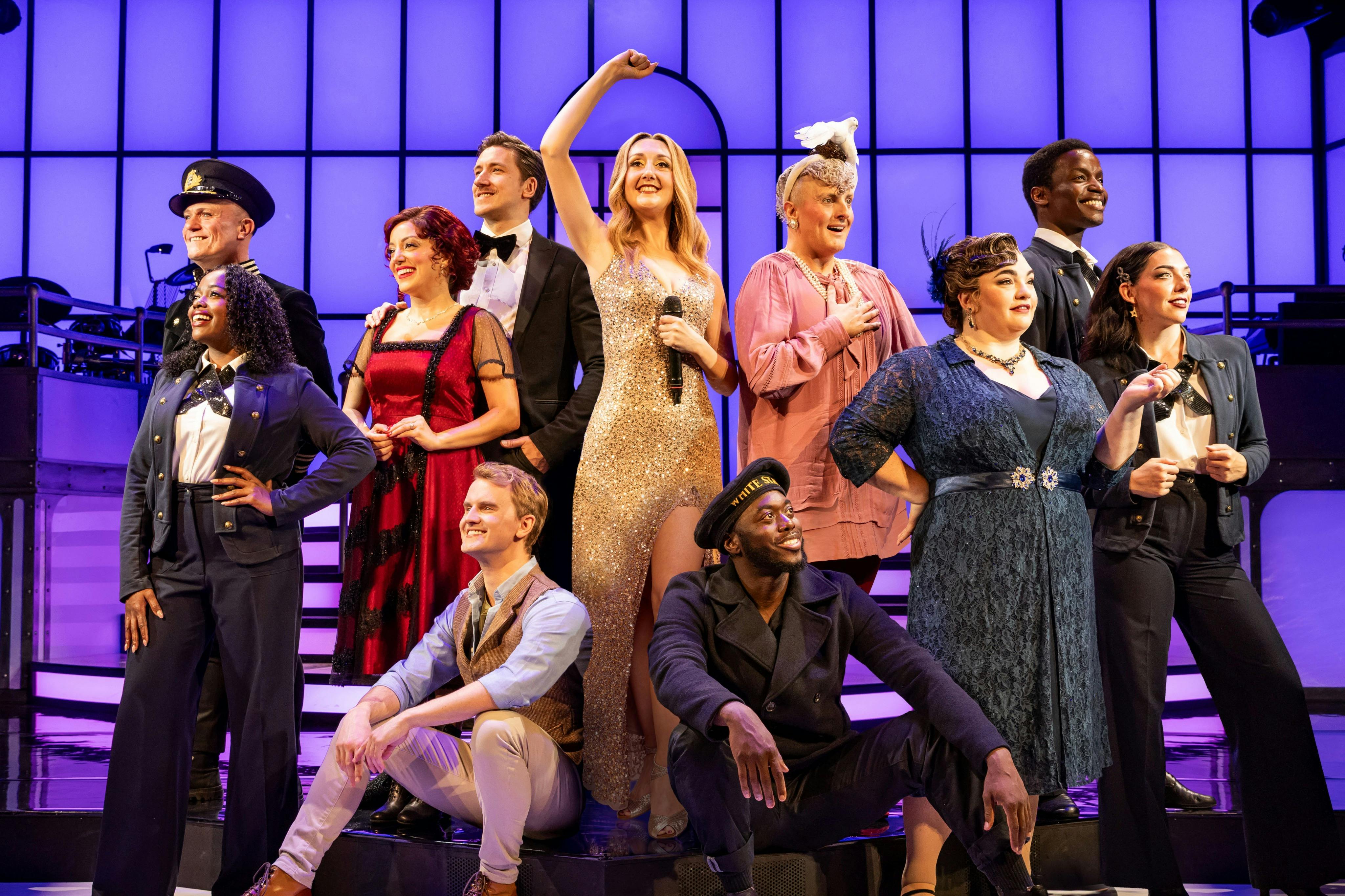 A group of people on stage dressed in various colorful costumes, smiling and posing under bright lights. A woman in the center holds a microphone.