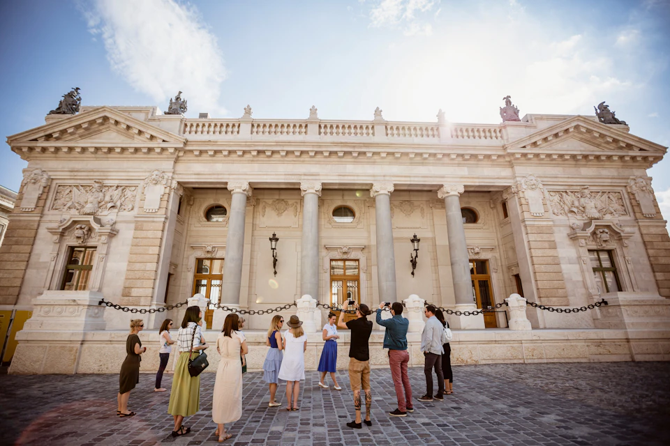 A group of people standing on a cobblestone square, observing and photographing a historic building with columns.