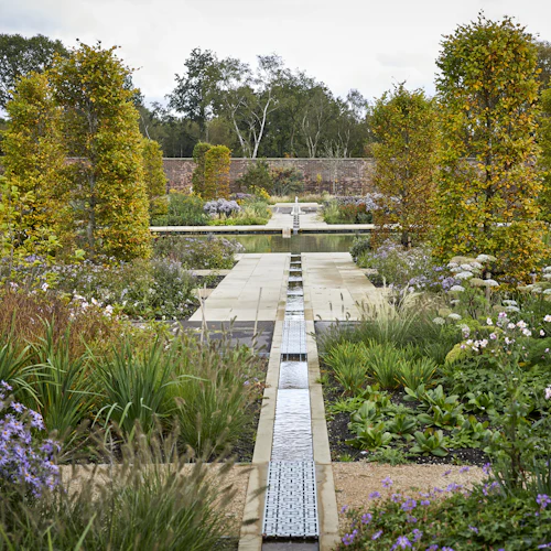 En formell trädgård med en central stenväg, kantad med trimmade träd, färgglada blommor och en vattenfunktion som löper genom mitten.