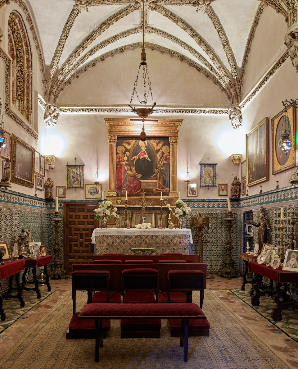 Small chapel with an ornate altar, religious paintings, and six red chairs. Chandeliers hang from the arched ceiling.