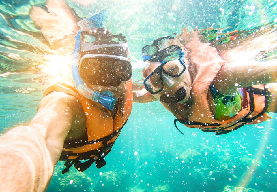 Two people snorkeling underwater, wearing orange life vests and masks, with sunlight filtering through the blue-green water.