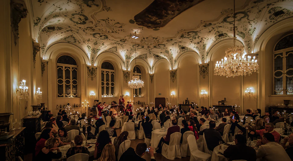 A formal dinner event in an opulent hall with high arched ceilings, chandeliers, and a string quartet performing to seated guests.
