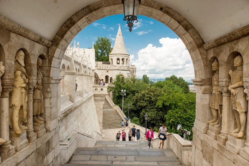 Stone archway framing people walking up steps towards Fisherman's Bastion, with trees and a blue sky with clouds visible.